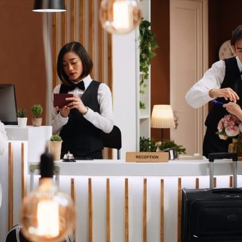 Tourists going through check in process at hotel reception desk, staff verifying passport id for registration. Two women signing ledger and reservation papers for accommodation on trip.